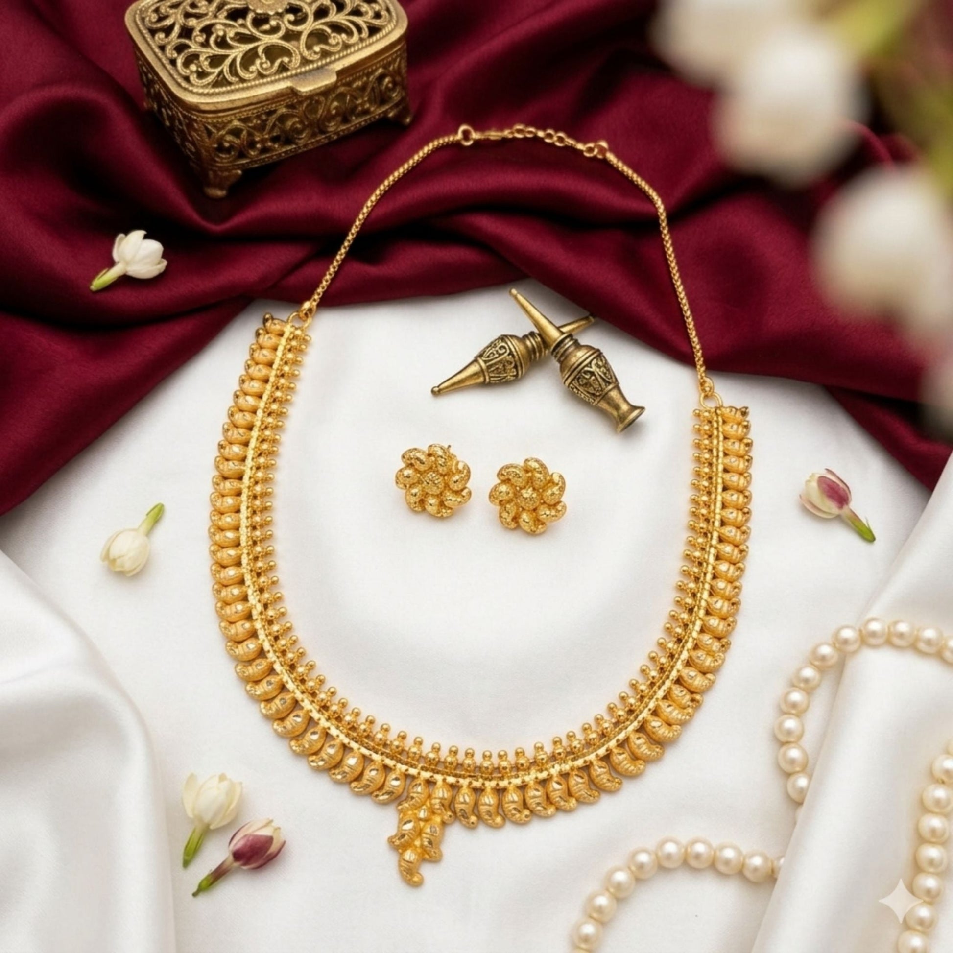 A gold-toned traditional necklace set with matching flower-shaped earrings, arranged on white satin with deep red fabric, pearl strands, and small white flowers. A decorative brass box is partially visible in the upper left.
