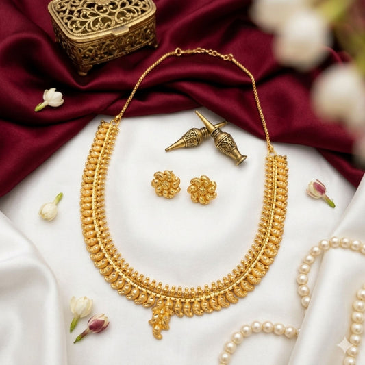 A gold-toned traditional necklace set with matching flower-shaped earrings, arranged on white satin with deep red fabric, pearl strands, and small white flowers. A decorative brass box is partially visible in the upper left.